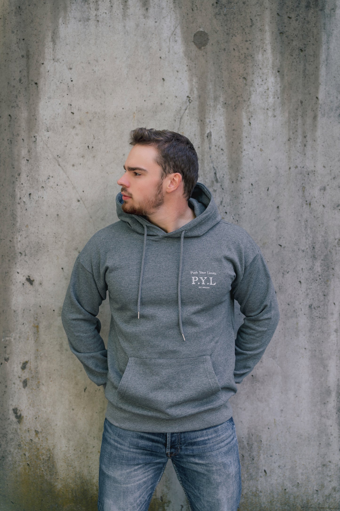 Man wearing a gray hoodie with 'P.Y.L' logo against a concrete wall