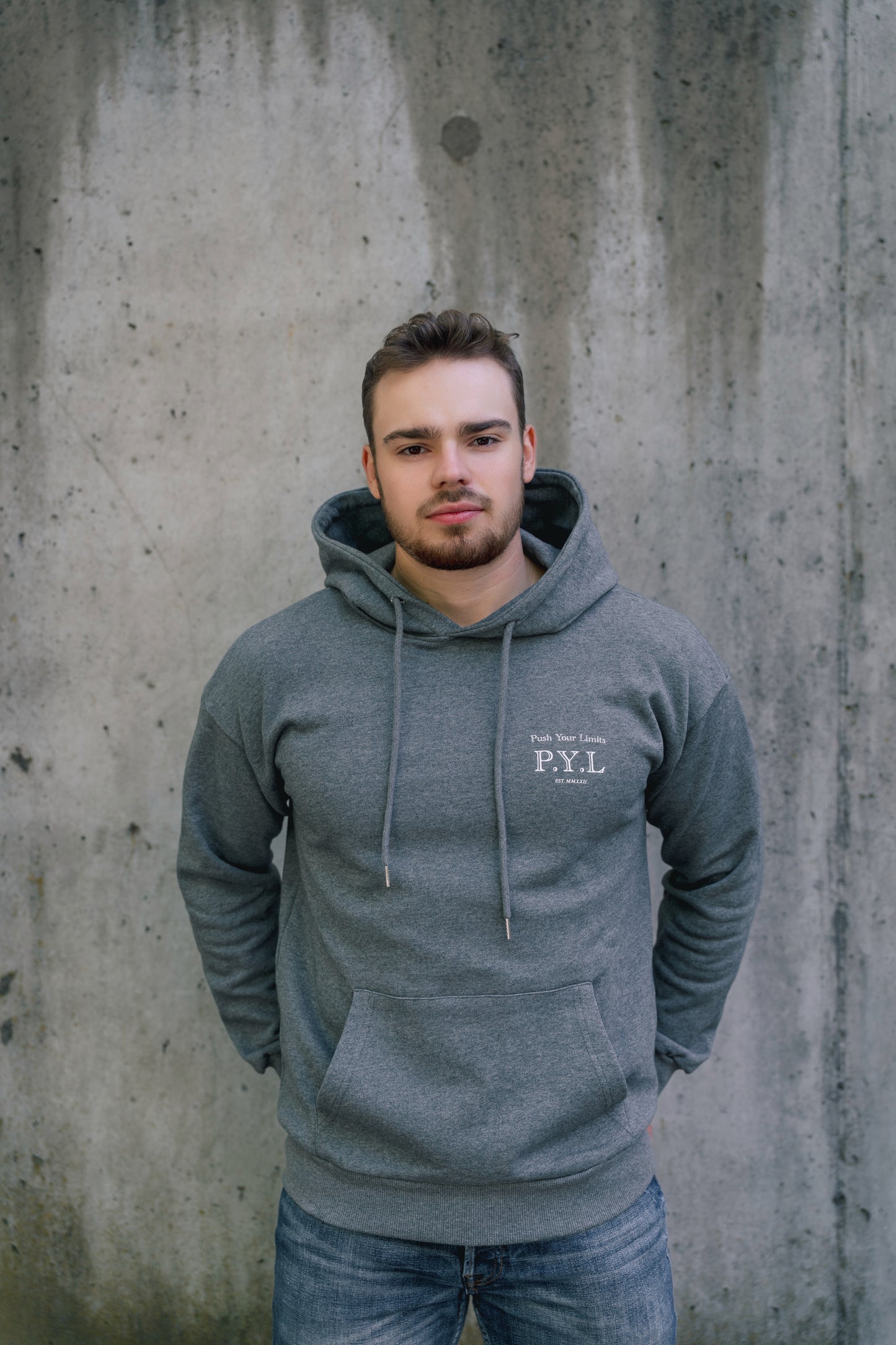 Man wearing a gray hoodie with 'P.Y.L' logo against a concrete wall