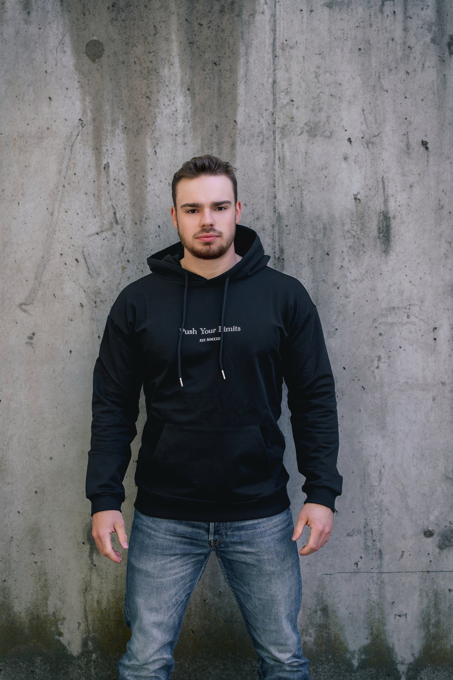 Man wearing a black hoodie with text standing against a concrete wall.