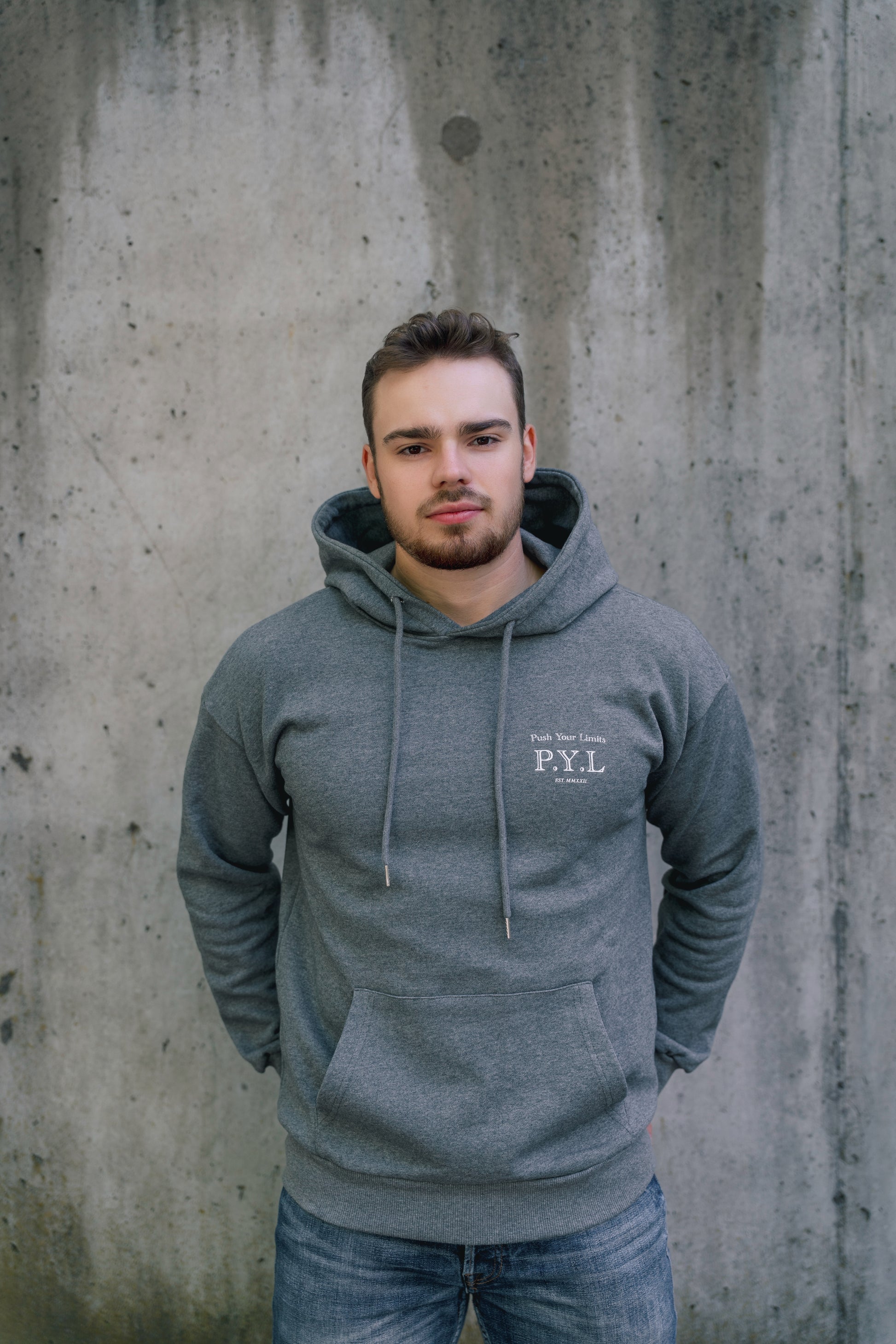 Man wearing a gray hoodie with 'P.Y.L' logo against a concrete wall